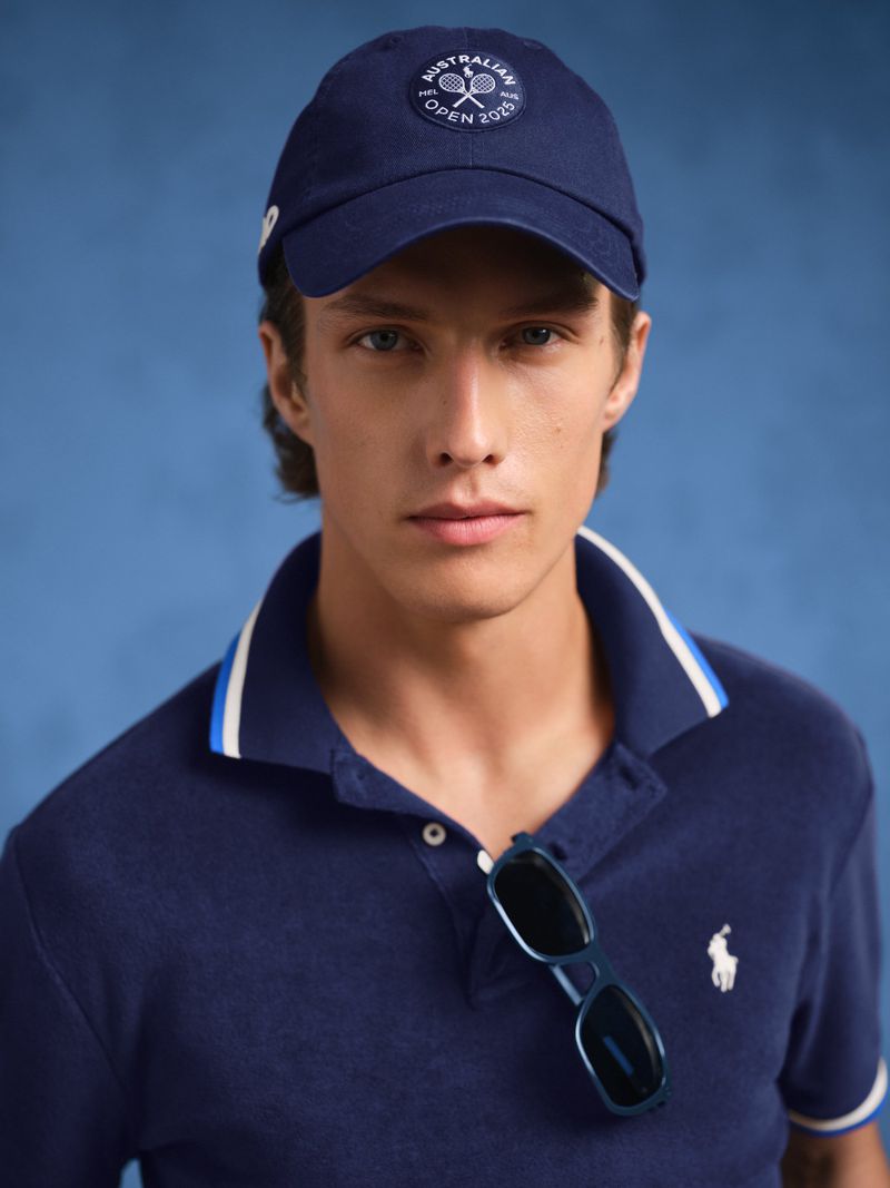 Close-up of an adult male wearing a blue polo shirt and matching baseball cap, sunglasses hanging from shirt. Blue background.