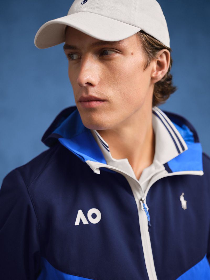 Man posing indoors wearing a baseball cap, zip-up jacket, and polo shirt, against a blue background, captured in a medium shot.