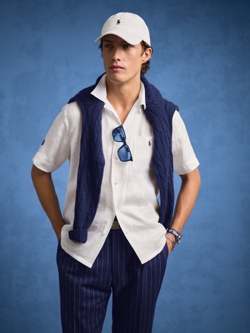 Man in a white shirt, navy sweater, and pinstripe pants, wearing a cap and sunglasses, poses against a blue background in a studio shot.