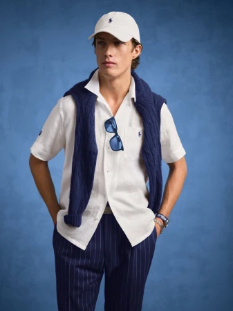 Man in a white shirt, navy sweater, and pinstripe pants, wearing a cap and sunglasses, poses against a blue background in a studio shot.