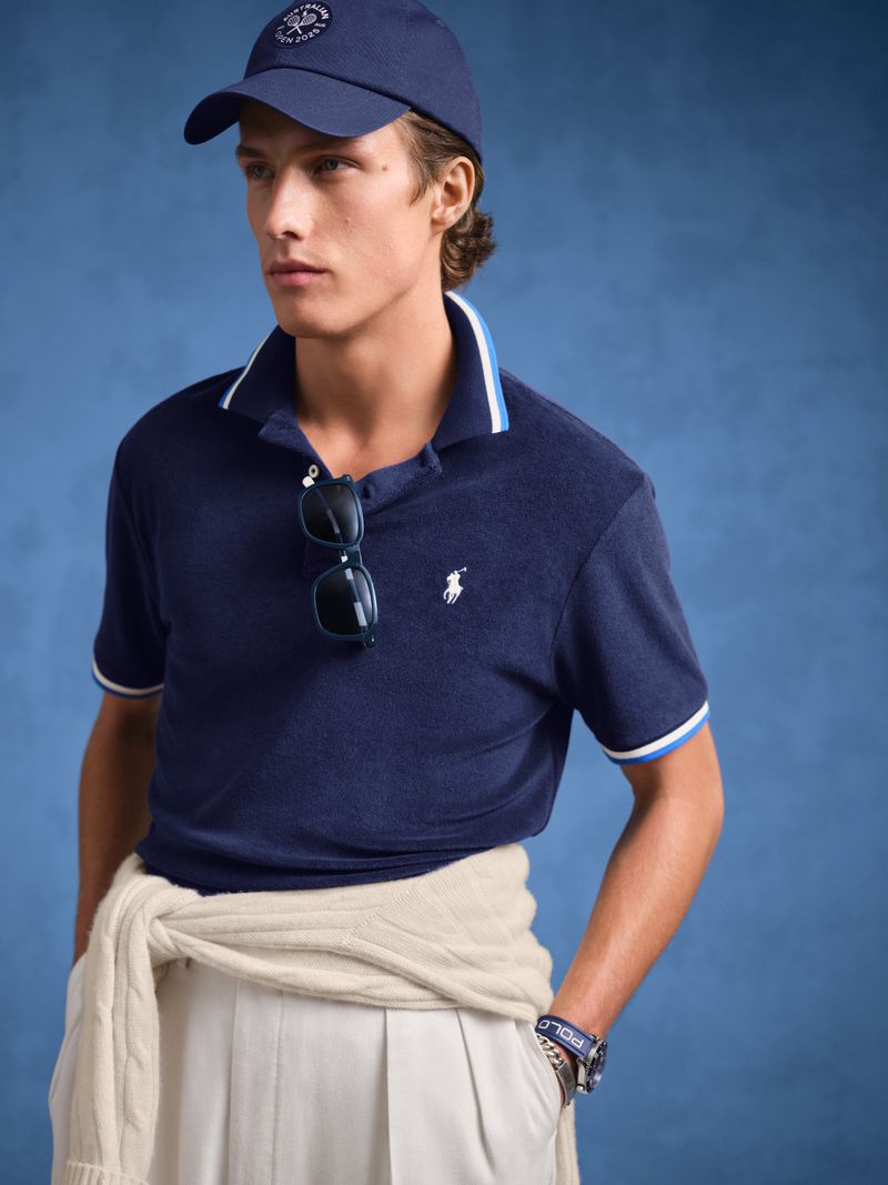 A young man wears a navy polo shirt, cream-colored pants, and a matching baseball cap, posing indoors with a blue backdrop.
