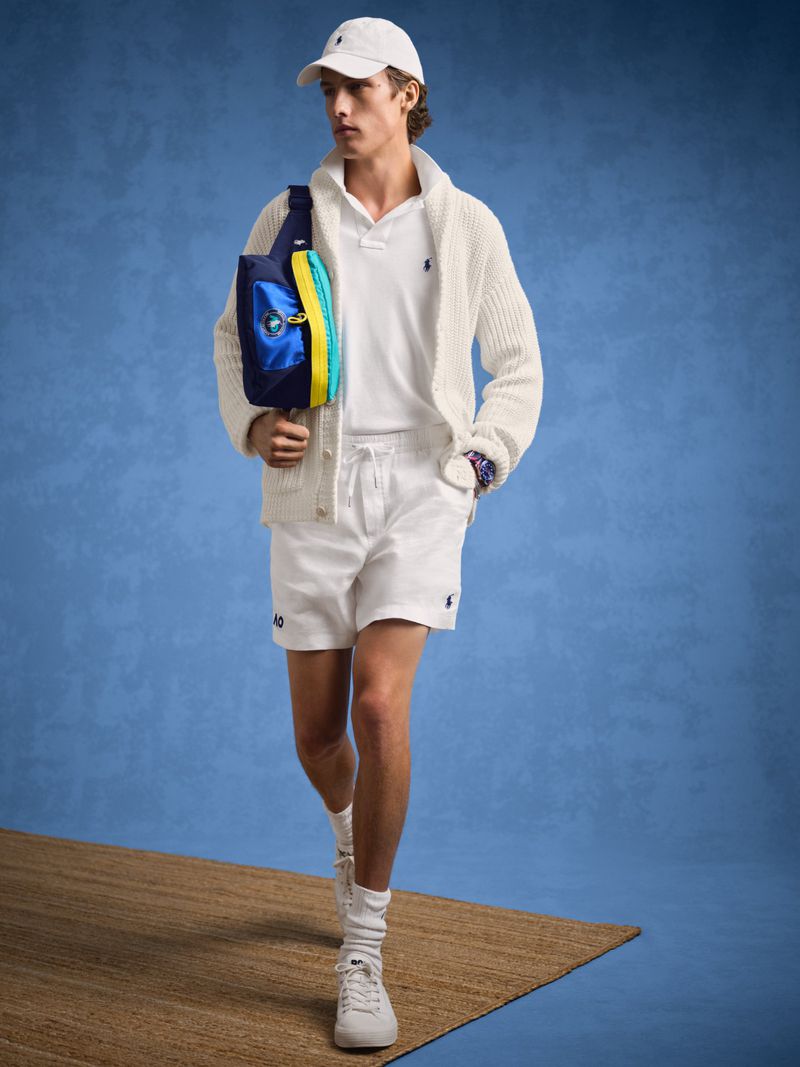 Man in white tennis outfit with shoulder bag walks on brown rug against blue background. Polo shirt, shorts, cardigan, cap, sneakers.