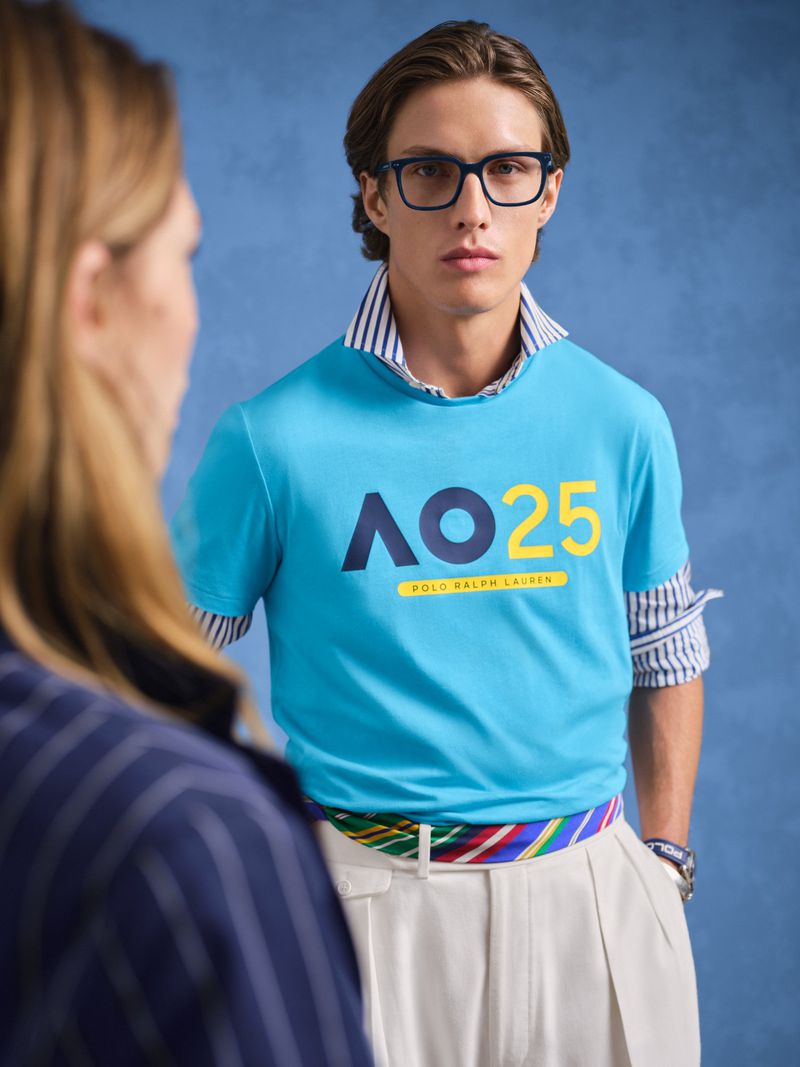 Man wearing a blue Polo Ralph Lauren shirt with the AO 25 logo, dress pants, and a striped belt stands indoors against a blue background.