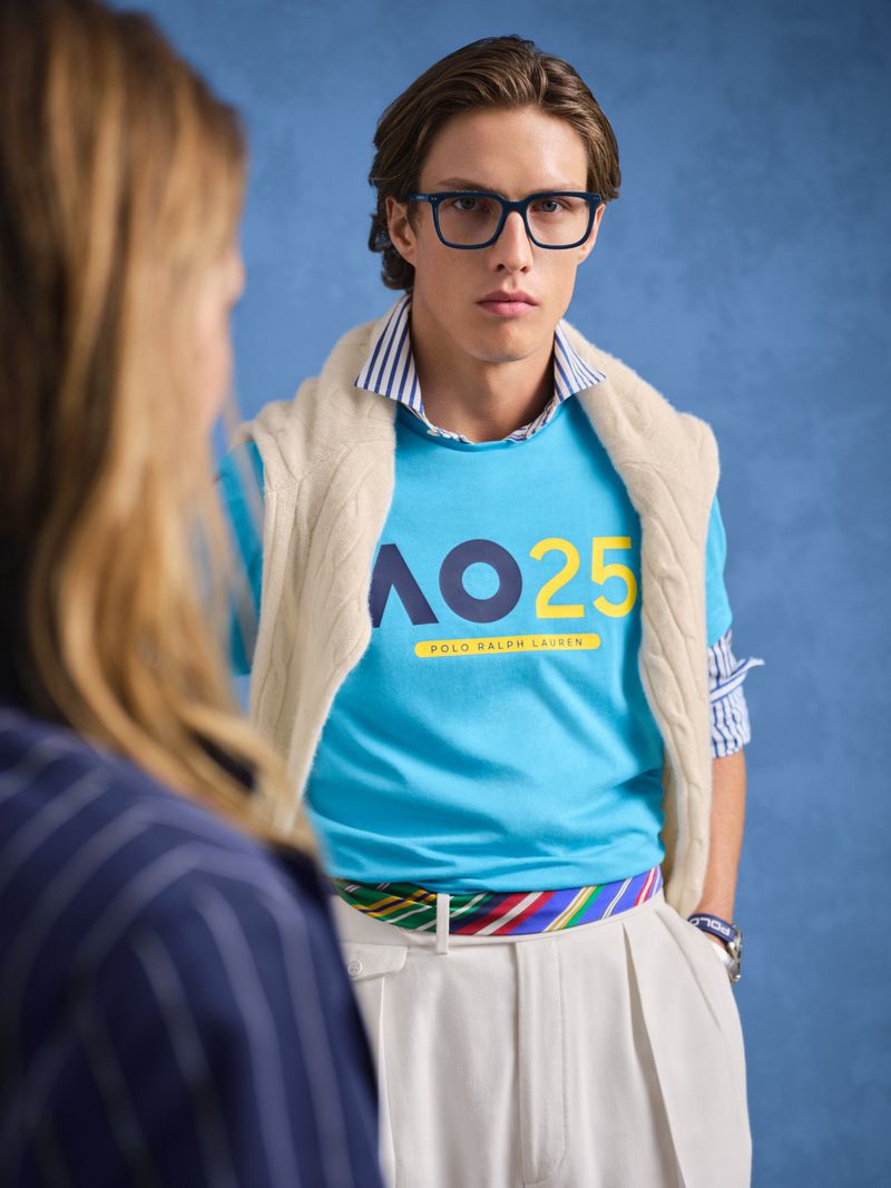 Stylish young adult male in turquoise logo tee, glasses, and preppy layered look posing with another person in a studio setting.