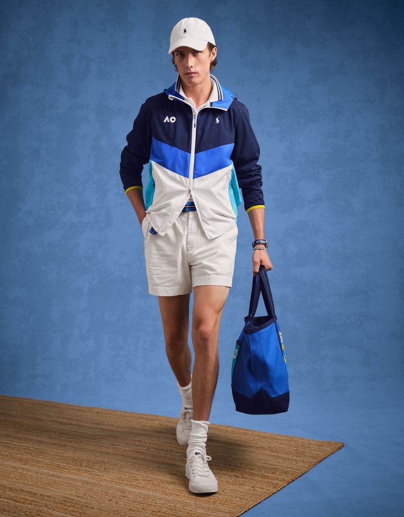 A fashionable adult male wearing a Ralph Lauren color-blocked jacket, shorts, cap and sneakers with a tennis bag against a blue background.