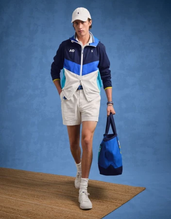 A fashionable adult male wearing a Ralph Lauren color-blocked jacket, shorts, cap and sneakers with a tennis bag against a blue background.