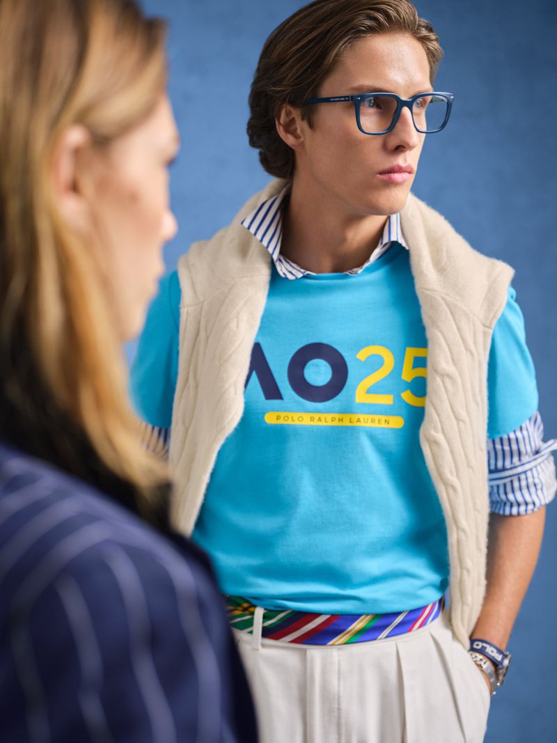 A young man in glasses wears a teal shirt with logo, white pants, and a colorful belt, next to a woman in a pinstripe jacket.