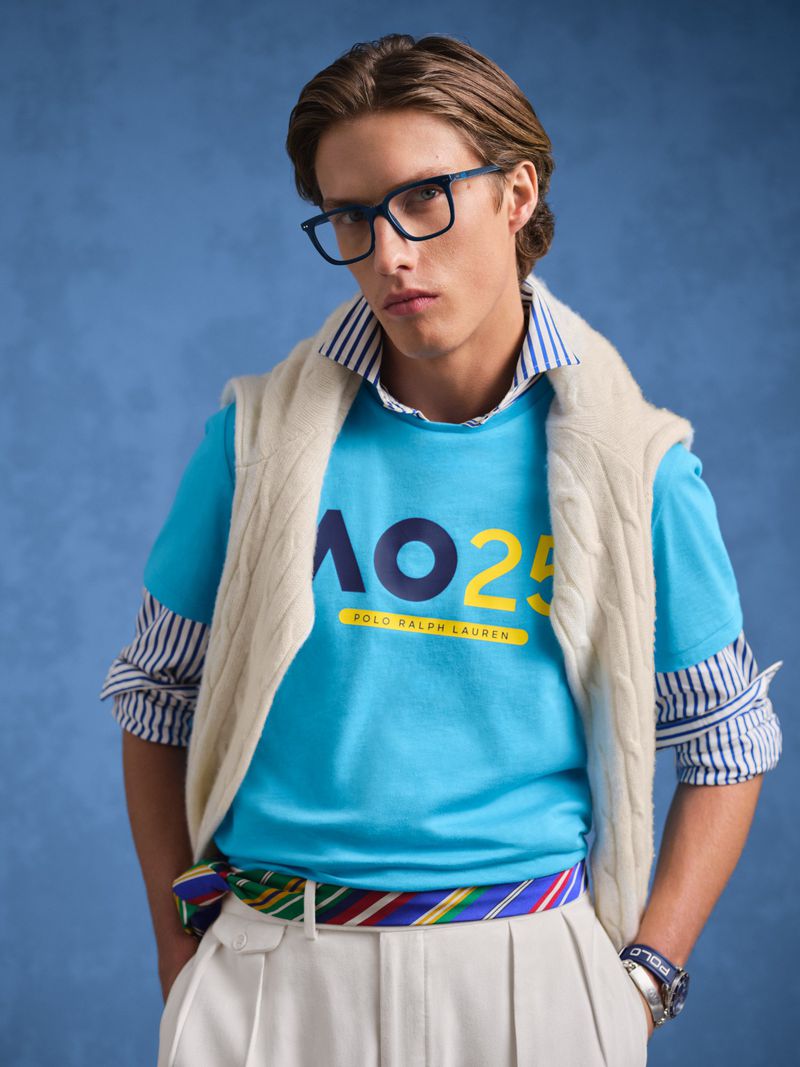 A fair-skinned adult male poses in a blue t-shirt with a logo and is accessorized with glasses, a sweater vest, and a striped belt, against a blue backdrop.