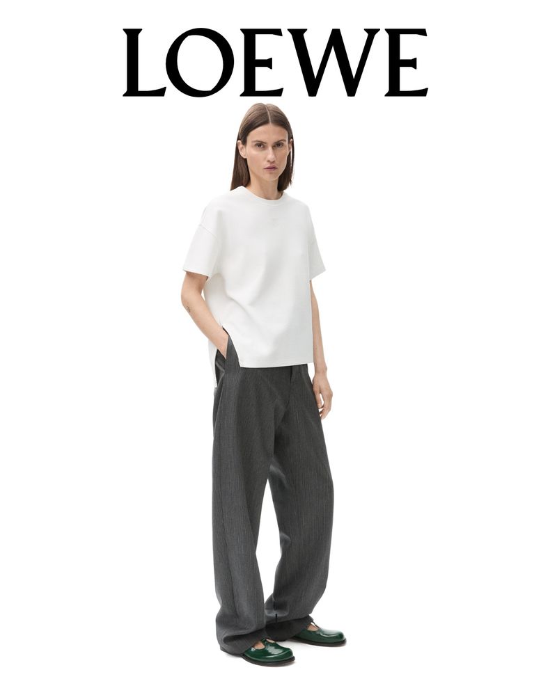 A woman models a white t-shirt and grey pants in a fashion studio shot against a white backdrop, showcasing a minimalist style.