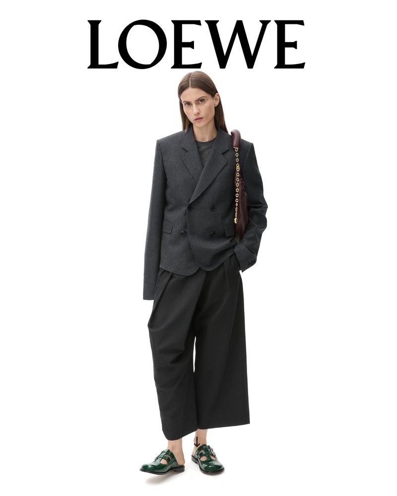 Fashionable woman in a grey blazer and dark culottes poses in a studio. Loewe logo at the top. Dark green sandal shoes.