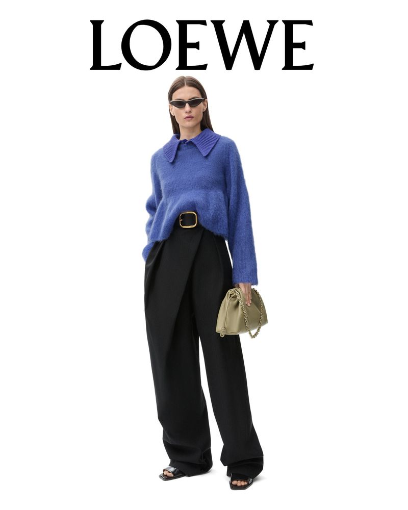 Model wearing dark sunglasses, blue textured sweater, and black trousers holding a beige handbag against a plain white background