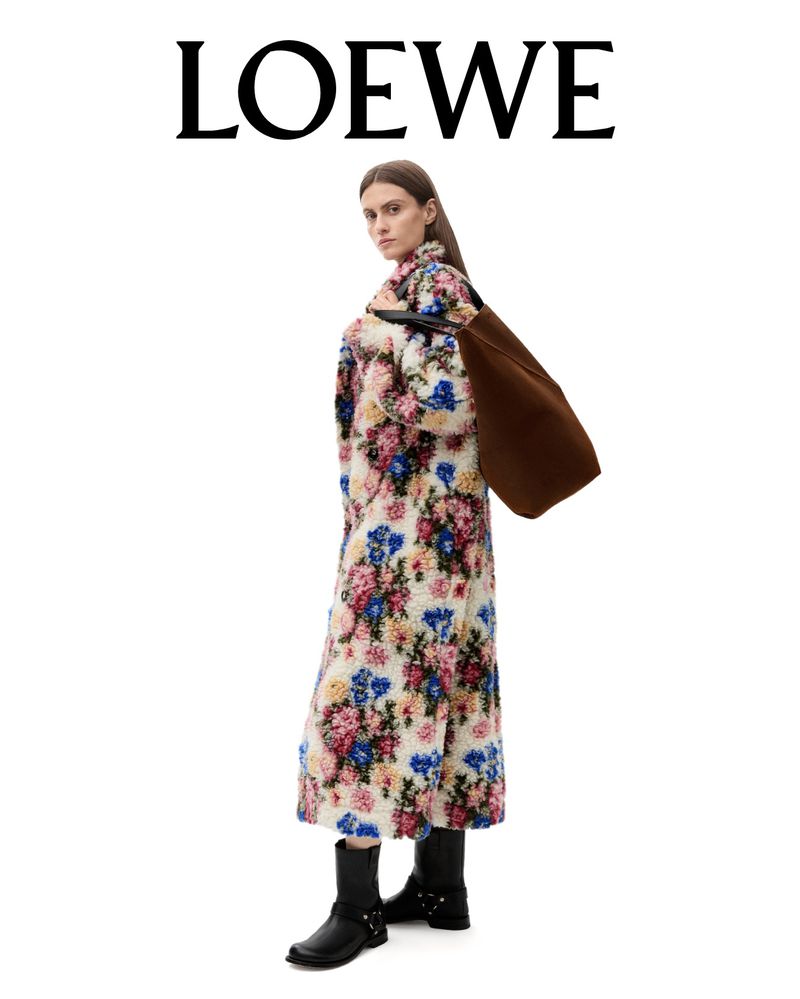 A woman models a long floral coat by LOEWE, standing in a white studio with a brown bag and black boots, fashion photography.