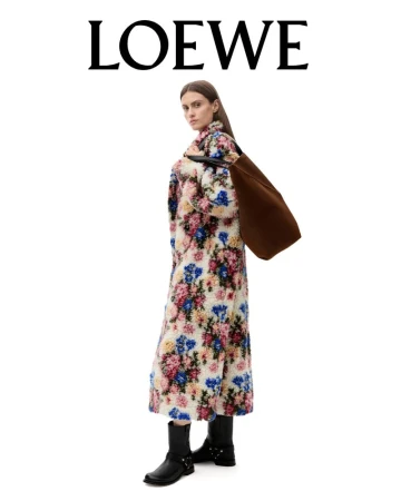 A woman models a long floral coat by LOEWE, standing in a white studio with a brown bag and black boots, fashion photography.
