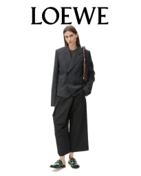 Model standing against a white backdrop wearing a dark grey blazer and black trousers holding a dark handbag.