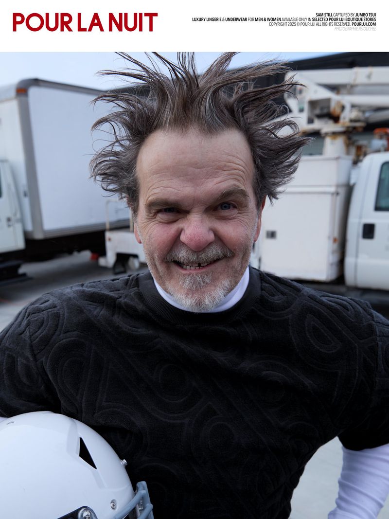 Portrait of a grinning man with spiky grey hair, wearing a patterned black shirt and holding a football helmet outdoors.
