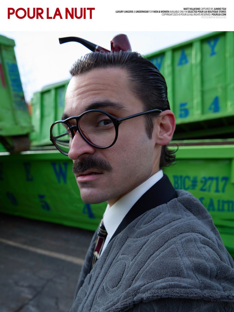 Portrait of a man with glasses and mustache, wearing a gray patterned jacket with a pipe balanced on his head, in front of green trailers.