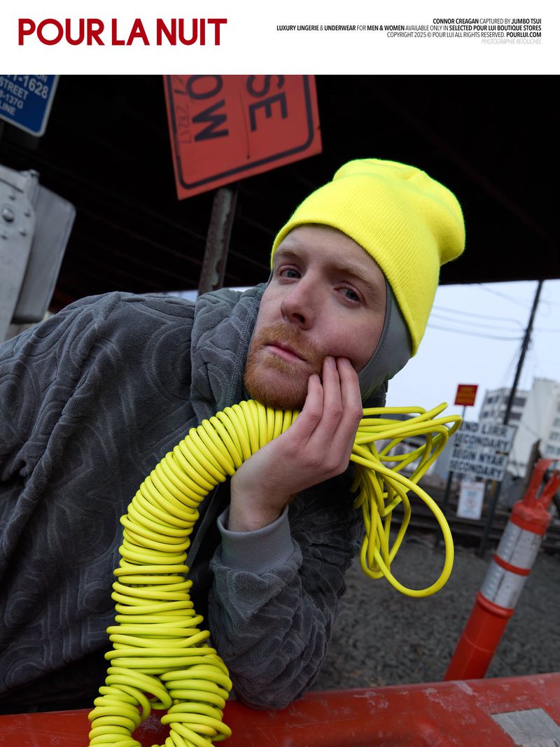 A man in a yellow beanie and gray hooded sweatshirt reclines against a red barrier with a yellow cable draped around his neck.