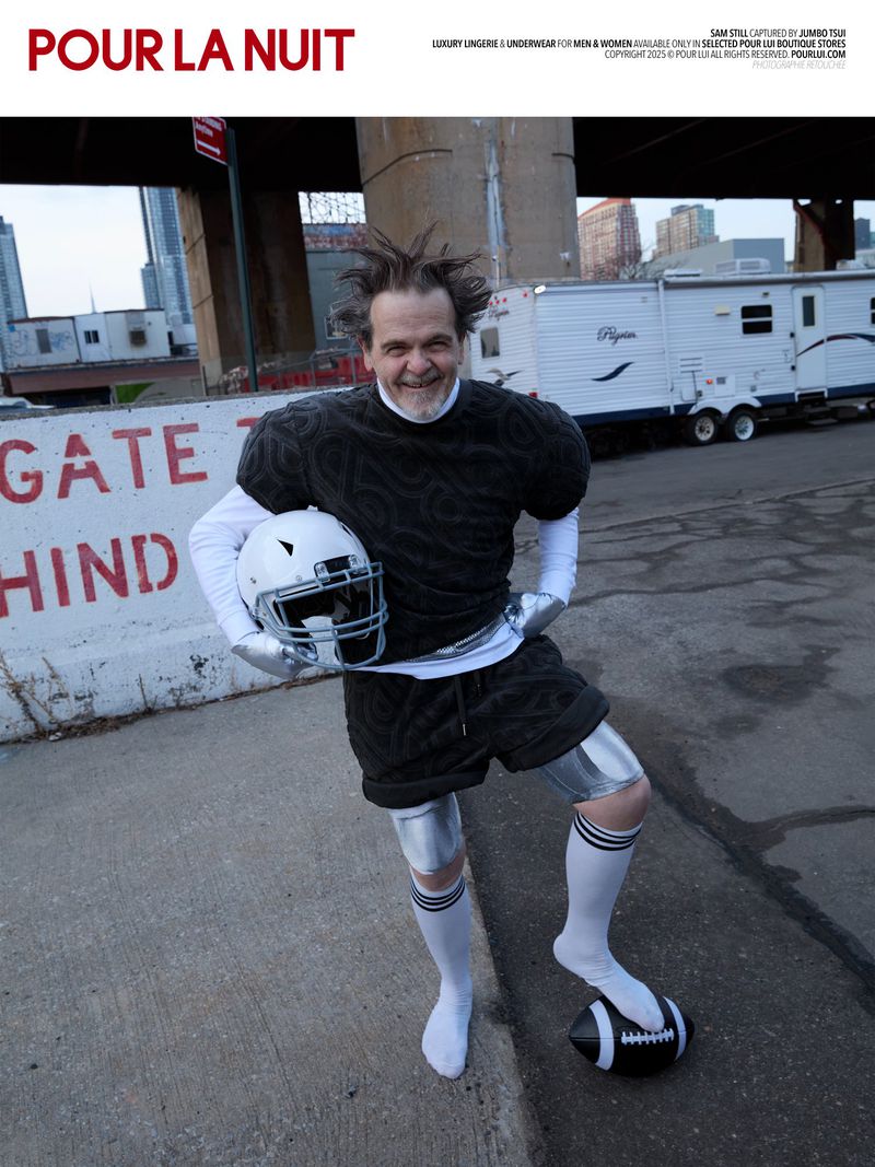 A man in football gear poses in an urban setting with a helmet and ball, with a trailer and bridge in the background.