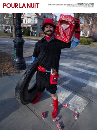 Man in red hat poses with tire and shopping bag near buildings and street lamp with soda cans scattered on sidewalk.