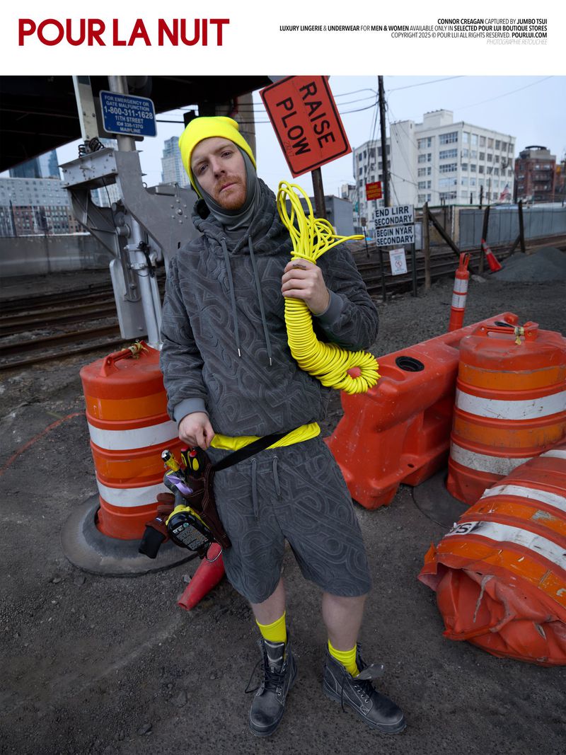Man in grey patterned hoodie and shorts, yellow beanie, holds yellow cable. Orange construction barrels and city backdrop.