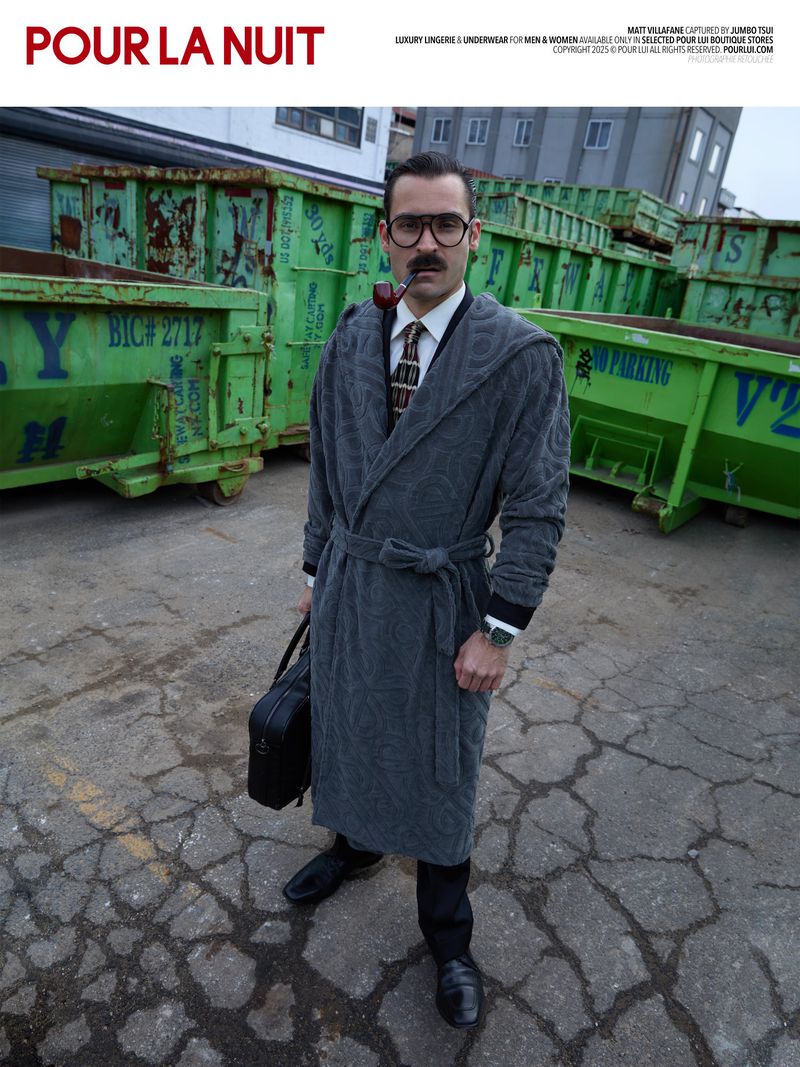 Man in gray bathrobe poses with pipe and briefcase in urban setting with dumpsters. Fashion-forward editorial photography.