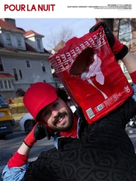 Smiling bearded man in a red cap and patterned black sweater holding a torn red soda can pack on his shoulder outdoors.