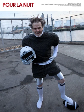 Man in padded black knitwear and athletic shorts posing with a football helmet and football on concrete by an industrial fence.