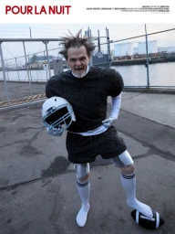 Man in padded black knitwear and athletic shorts posing with a football helmet and football on concrete by an industrial fence.