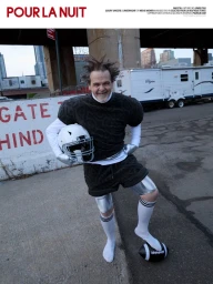 Man wearing a textured dark short-sleeved top and shorts, holding a white football helmet outdoors near a highway overpass.