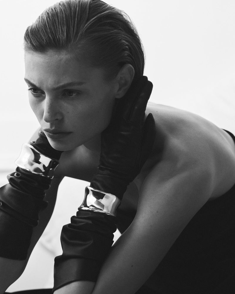 Black and white portrait of a person with slicked-back hair wearing black gloves with silver wristbands, against a bright white backdrop.