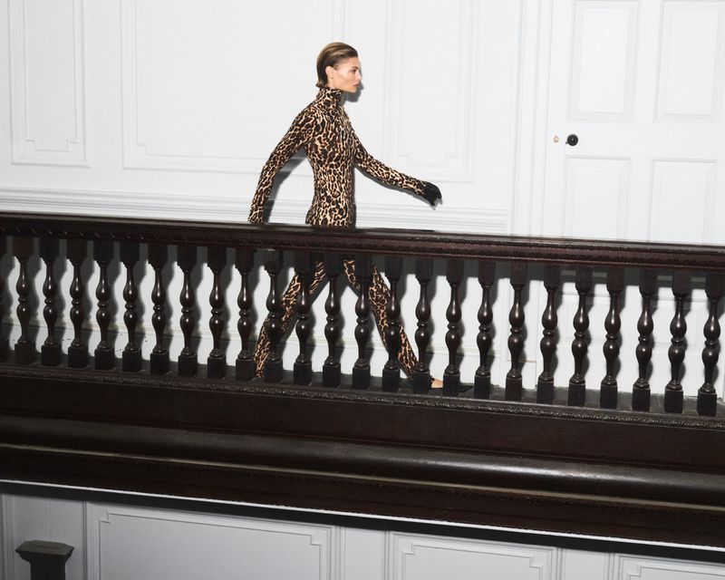 A slender woman in a leopard-print catsuit confidently walks indoors along a landing with dark wood railings and a white paneled wall.