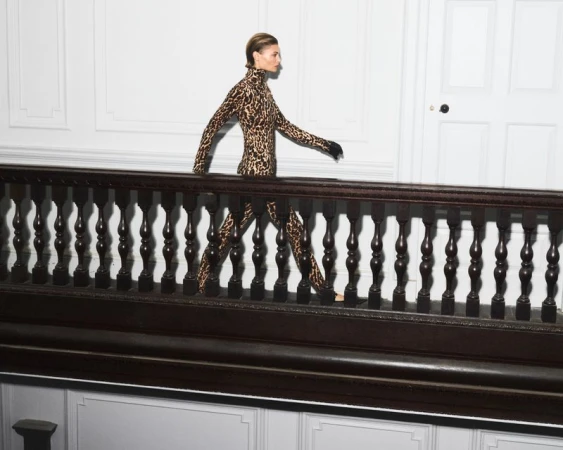A slender woman in a leopard-print catsuit confidently walks indoors along a landing with dark wood railings and a white paneled wall.