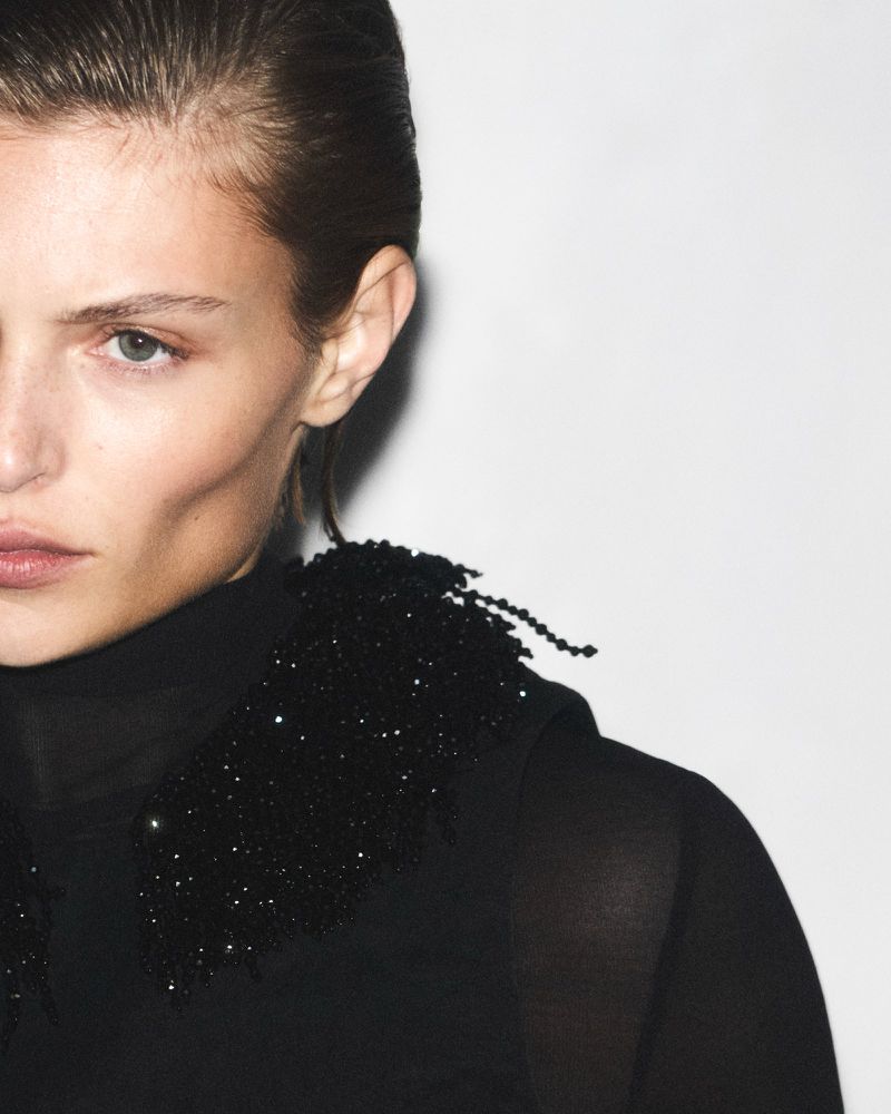 Close-up of a fair-skinned woman with a dark collared top, gazing towards the camera against a minimalist white backdrop.