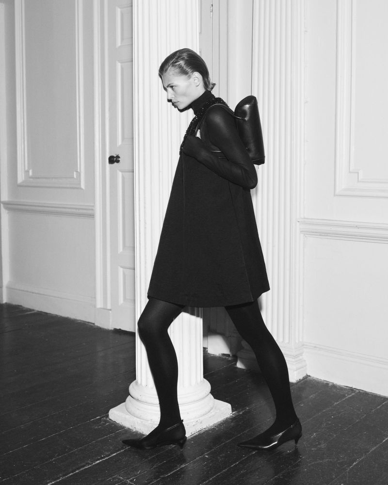 Black-and-white fashion shot of a female model in a black dress and black tights, carrying a rounded black backpack in an indoor setting.