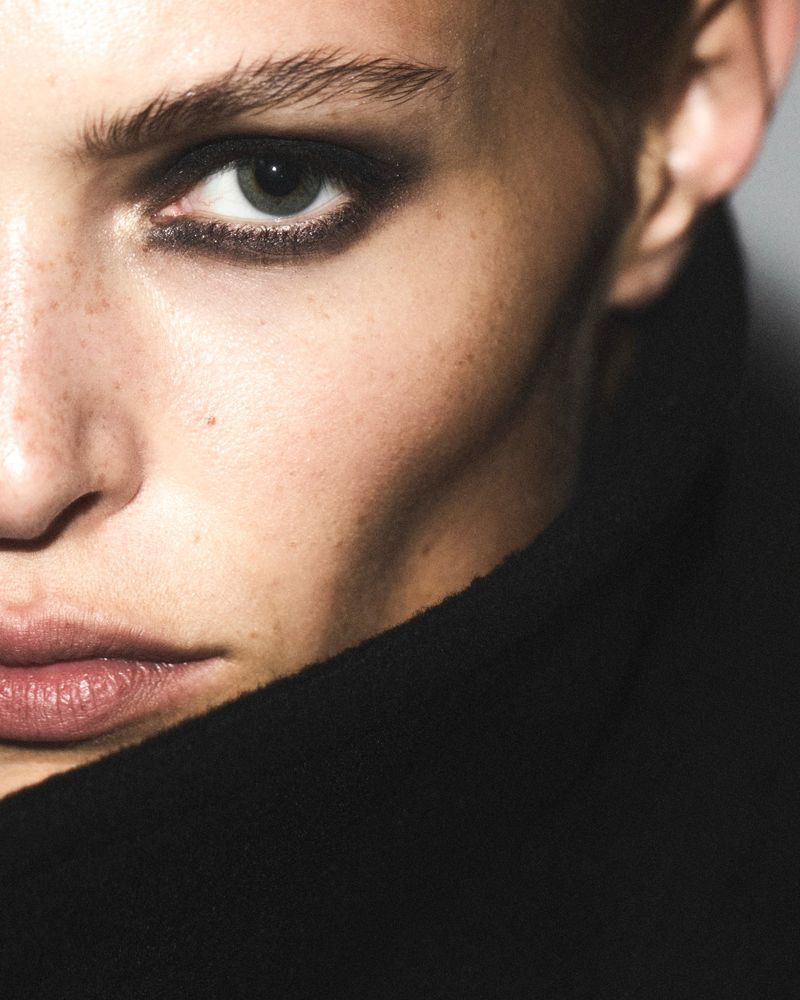 Close-up portrait of a fair-skinned adult woman with dark eyeshadow and a neutral expression, wearing a black high-neck garment against a grey background.