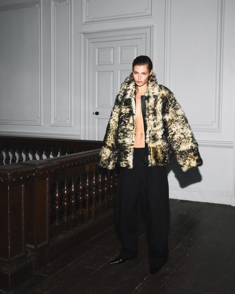 Model Bibi Breslin wearing a mottled faux-fur jacket and wide-leg trousers standing on a landing beside a wooden staircase banister.