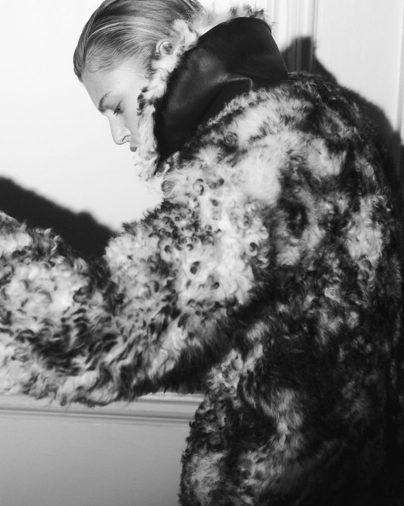 Monochrome shot of a woman in profile wearing a patterned fur coat with a dark collar, set against a plain light-colored wall.