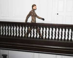 Model in leopard print jumpsuit walking past a dark wooden staircase railing indoors