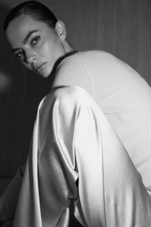 Black and white portrait of a model wearing a light-colored sweater and satin pants looking over her shoulder in a studio setting.