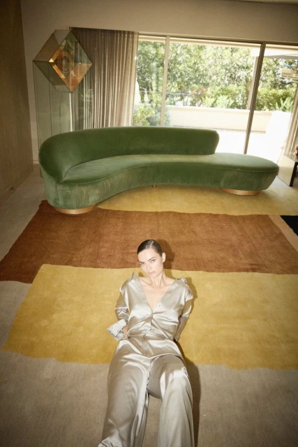 Adult female in silk loungewear reclines on a beige carpet in a modern interior with a green velvet couch near a large window.
