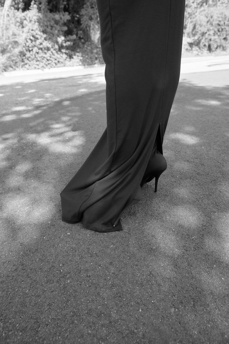 A black and white photo shows the lower portion of a person wearing a long black dress and high heels on a grassy surface with dappled shadows.