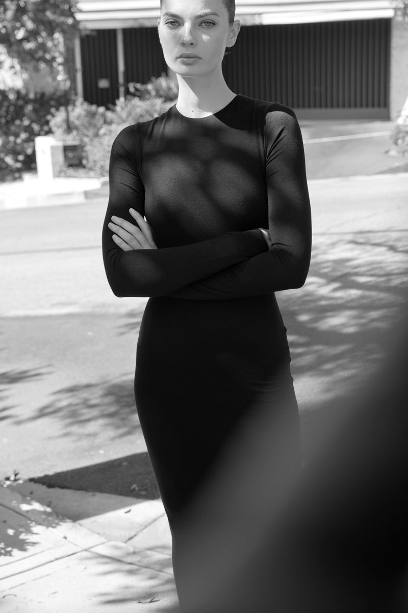 A slender adult woman in a long-sleeved black dress poses with arms crossed outdoors on a sunny day with tree shadows.