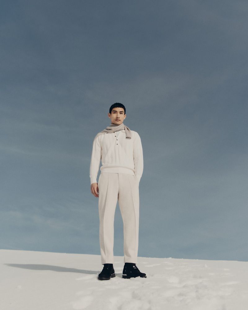 Fashion shot of a full-length male model wearing a cream-colored knit sweater, beige pants, a grey scarf, and black boots, outdoors.