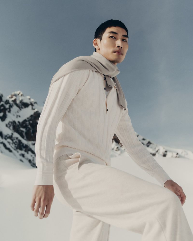 A fashionable Asian male model poses in an all-white outfit and scarf against a snowy mountain backdrop on a sunny day.