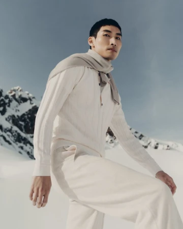 A fashionable Asian male model poses in an all-white outfit and scarf against a snowy mountain backdrop on a sunny day.