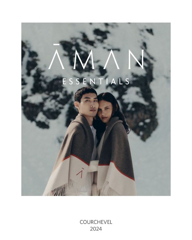 A couple, a man and a woman, are wrapped in blankets in a snowy environment with the brand logo 'AMAN ESSENTIALS' overhead.