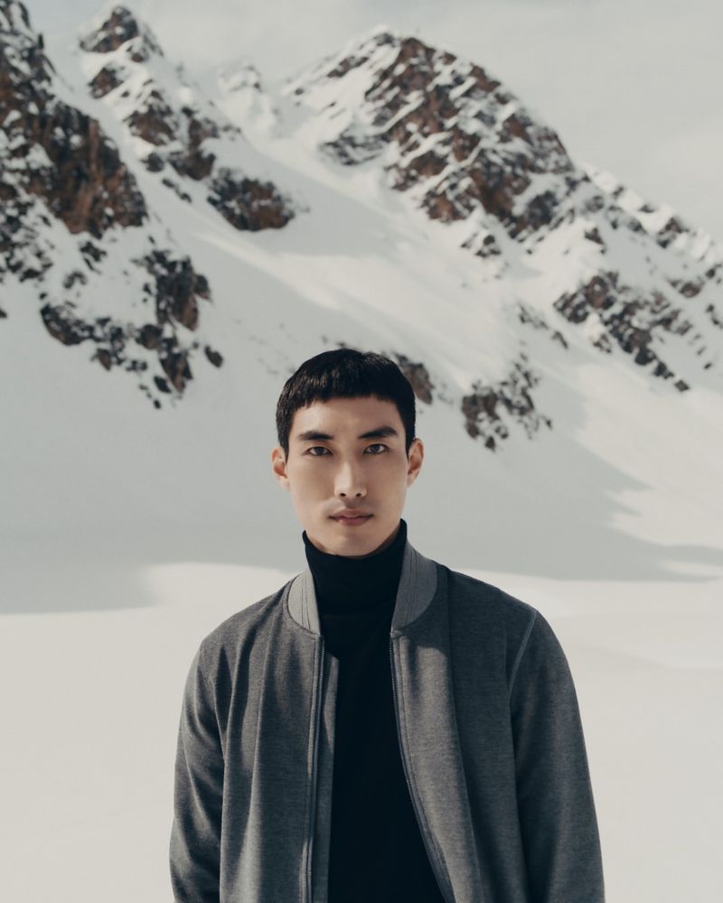 Adult male in a grey jacket and black turtleneck poses in a snowy, mountainous landscape on a winter day, medium shot.