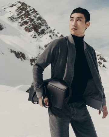 Asian man in a gray jacket and black turtleneck holding a black leather bag, posing against a backdrop of snow-covered mountains.