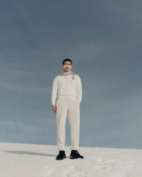 Model Do Byung Wook in a cream knit sweater, tailored trousers, and draped scarf standing on white sand dunes against a pale sky.