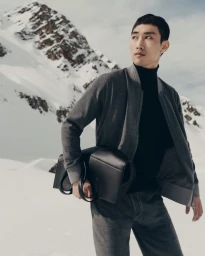 Asian male model in grey bomber jacket holding a black leather bag against a snowy mountain landscape